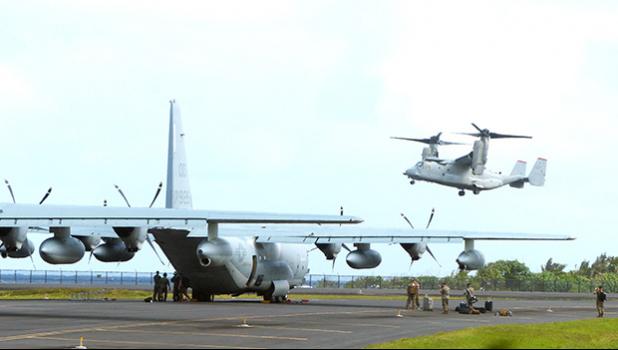 A C-130 on the tarmac and an MV-22 Osprey military plane overhead at Pago Pago International Airport