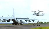 A C-130 on the tarmac and an MV-22 Osprey military plane overhead at Pago Pago International Airport