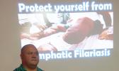 A Health Department official standing in front of a screen — in the background — showing a photo of a person affected with Lymphatic Filariasis